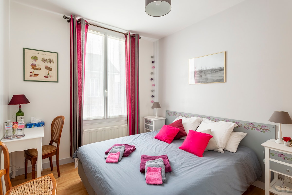 A cozy bedroom features a spacious bed adorned with colorful pillows and neatly arranged towels. Natural light filters through a large window framed by vibrant curtains. A small table and chair are positioned next to the bed, complemented by decorative wall art.