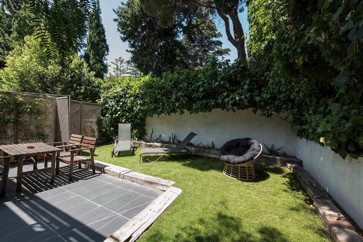 A spacious garden area is visible, featuring a green lawn surrounded by lush greenery. A picnic table with wooden chairs and a lounge chair are positioned on the patio. A circular, wicker lounge chair offers a cozy spot for relaxation, inviting peaceful outdoor moments.