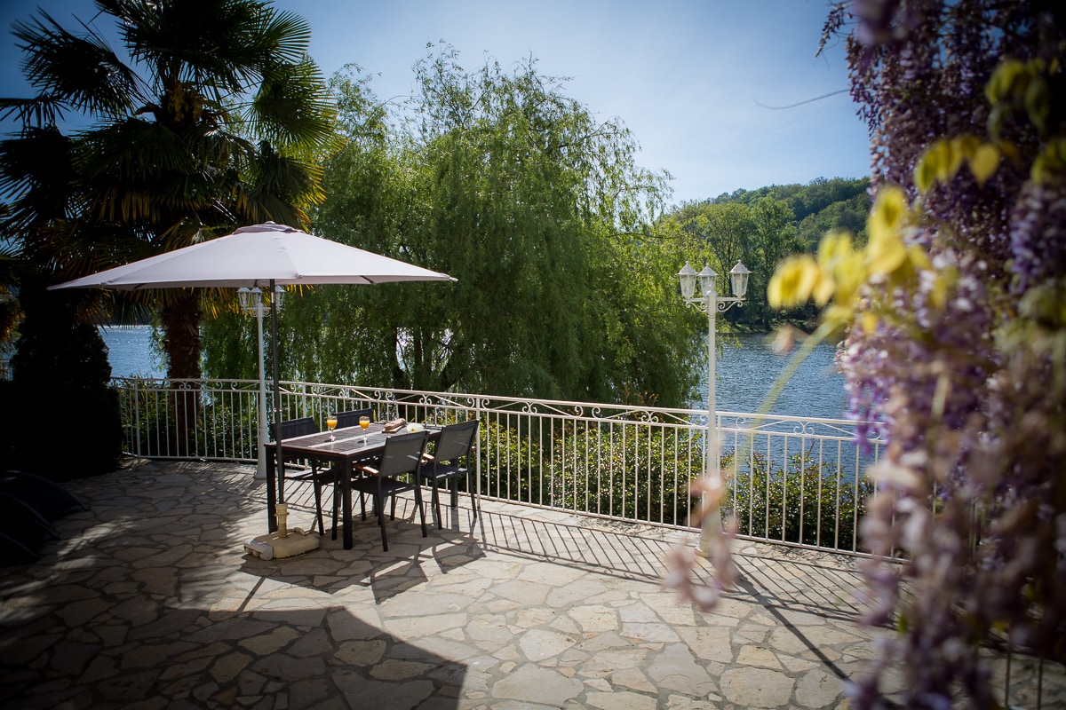 An outdoor terrace is visible, featuring a large table surrounded by chairs and shaded by a parasol. Lush greenery lines the space, with a view of the Dordogne River and distant hills beyond. Decorative plants add charm along the edge of the terrace.