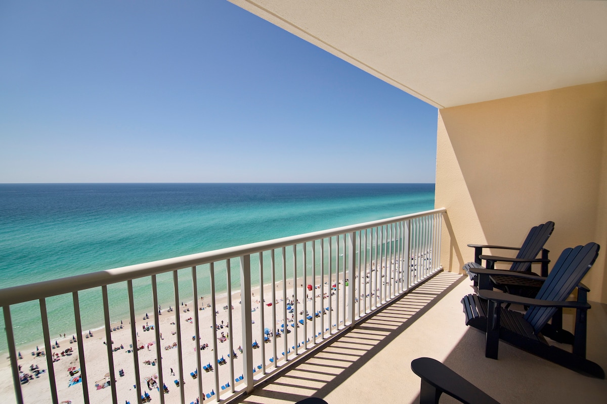 A private balcony offers stunning views of the clear blue ocean, with soft waves gently lapping at the white sandy beach below. Two comfortable chairs are positioned for relaxation, while the vast expanse of sky and water creates a serene atmosphere.