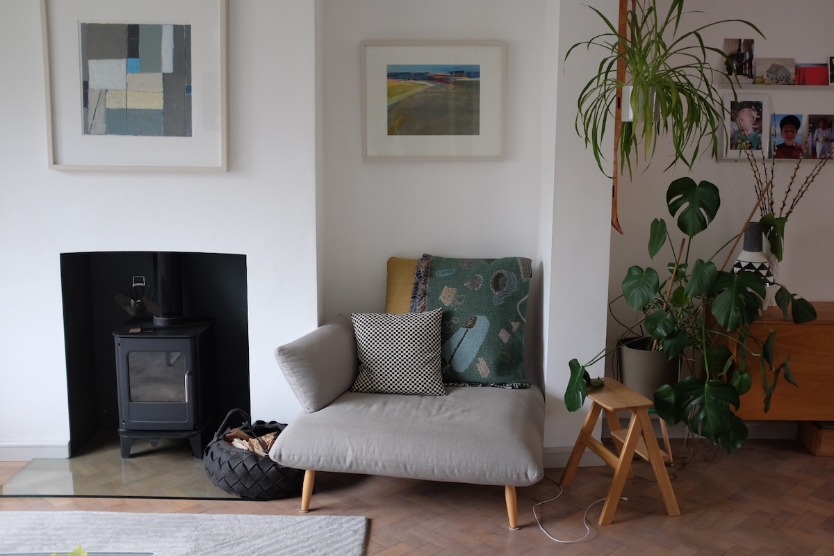 A cozy corner is shown, featuring a modern chair adorned with patterned cushions. A small wooden stool sits beside the chair, and a black wood stove is positioned against the wall. A green plant and framed artwork bring a touch of nature and color to the space.