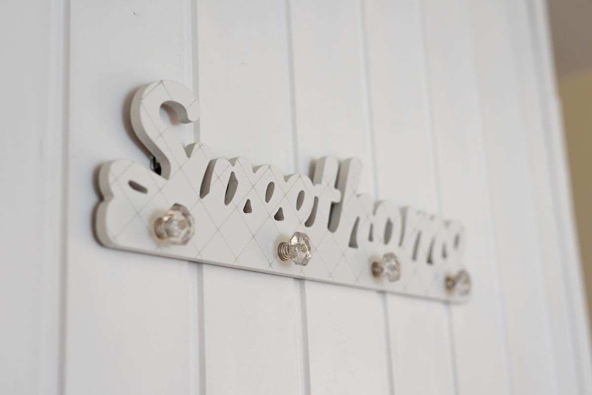 A decorative wall sign reading 'Sweet Home' is mounted on a white wooden surface. The sign features a stylish font and is enhanced by crystal-like knobs that add a touch of elegance.