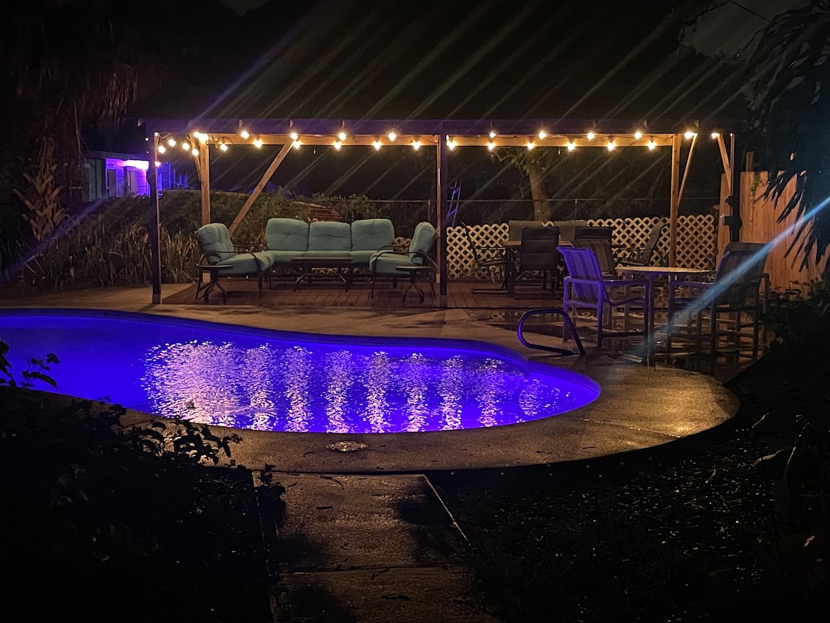 A serene outdoor space features a night-lit pool reflecting soft blue hues. Comfortable seating is arranged under a pergola, adorned with string lights, creating an inviting atmosphere for relaxation. Lush greenery surrounds the area, enhancing the tranquil setting.