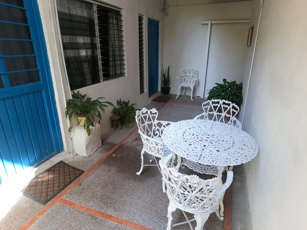 A shared patio features white, ornate outdoor furniture including a round table and four chairs. Lush green plants are positioned against the walls, adding touches of nature. The entrance is marked by a vibrant blue door, creating a contrast with the neutral-toned walls.
