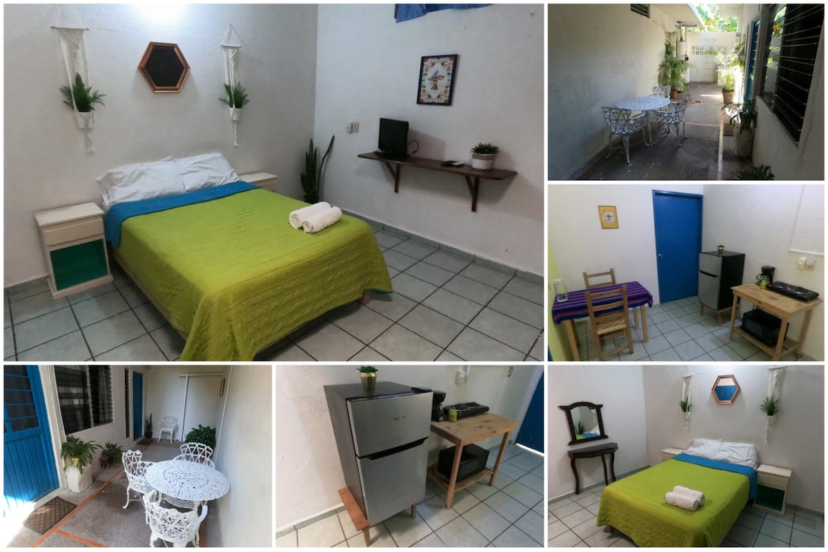 The room features a spacious bed covered with a bright green blanket and towels neatly arranged. A wall-mounted shelf and a small table are visible, along with plants adding a touch of greenery. A compact kitchenette with a refrigerator is also present, enhancing convenience.