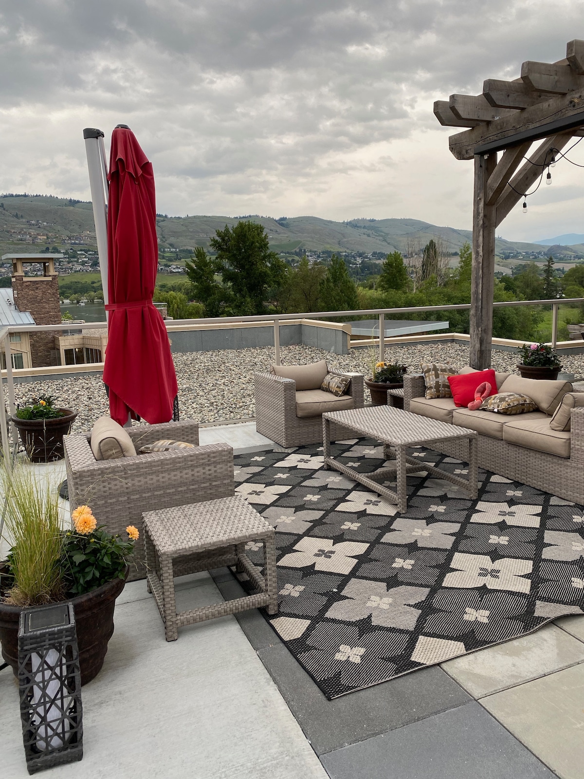 A rooftop patio is showcased with comfortable seating arrangements including a sectional sofa and side tables. A large umbrella provides shade while potted plants add greenery. Scenic views of hills and landscape are visible in the background, enhancing the open atmosphere.