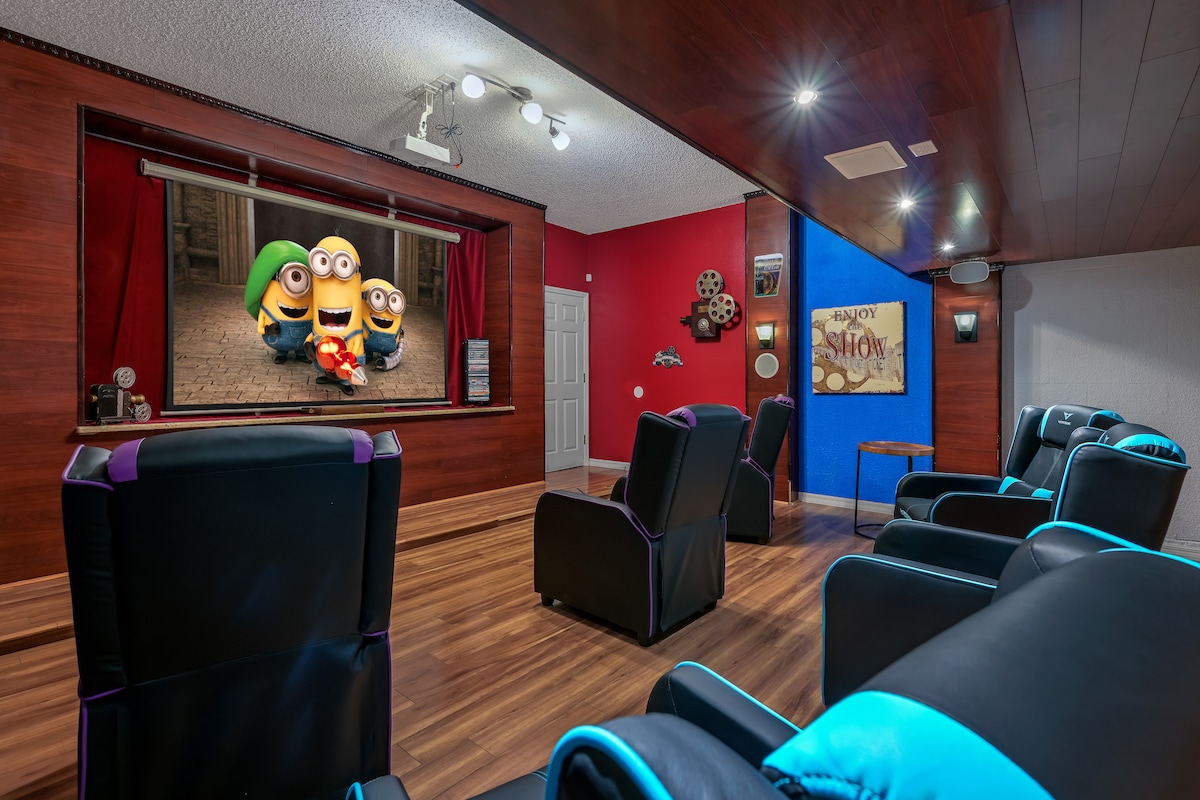 The movie theater features a large 150-inch screen with a vivid projection of Minions, surrounded by plush seating arranged for comfort. The room exhibits a cozy ambiance, with dark wood paneling and colorful accents, creating a dedicated space for entertainment.