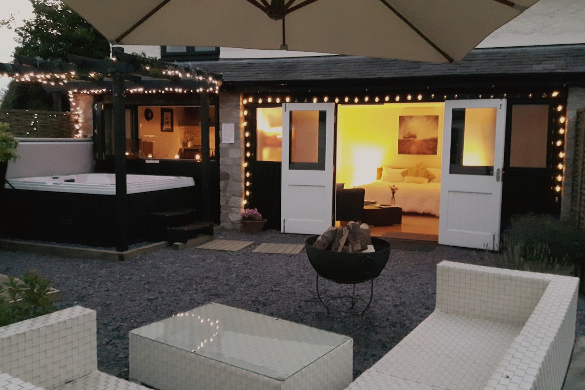 An outdoor area is presented with a hot tub, surrounded by soft lighting from strings of fairy lights. White outdoor furniture includes a coffee table and seating, providing a relaxed atmosphere. The entrance to the cozy interior is visible, showcasing warm lighting that illuminates a spacious bedroom.