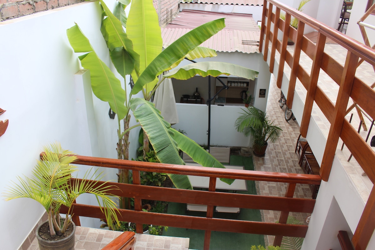 An open-air courtyard is surrounded by lush greenery and decorative plants. A wooden railing adds a rustic touch while offering views of comfortable seating areas below, framed by tropical elements like tall banana plants. Sunlight streams into the space, enhancing its natural appeal.
