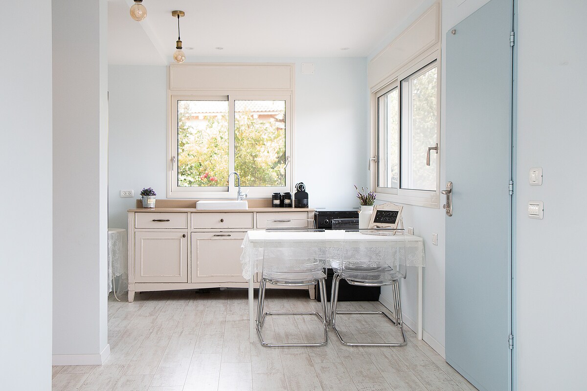 A well-organized kitchenette features light-colored cabinetry and a spacious countertop. Natural light enters through large windows, illuminating a small dining table with transparent chairs. Kitchen appliances, including a Nespresso machine, are neatly arranged, contributing to a functional cooking space.