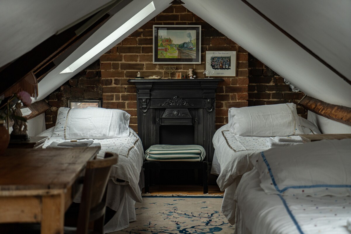An attic room features exposed brick walls and sloped ceilings, furnished with two spacious beds. A decorative fireplace serves as a focal point, while a wooden table and chairs occupy one corner. Art pieces decorate the walls, enhancing the cozy ambiance.