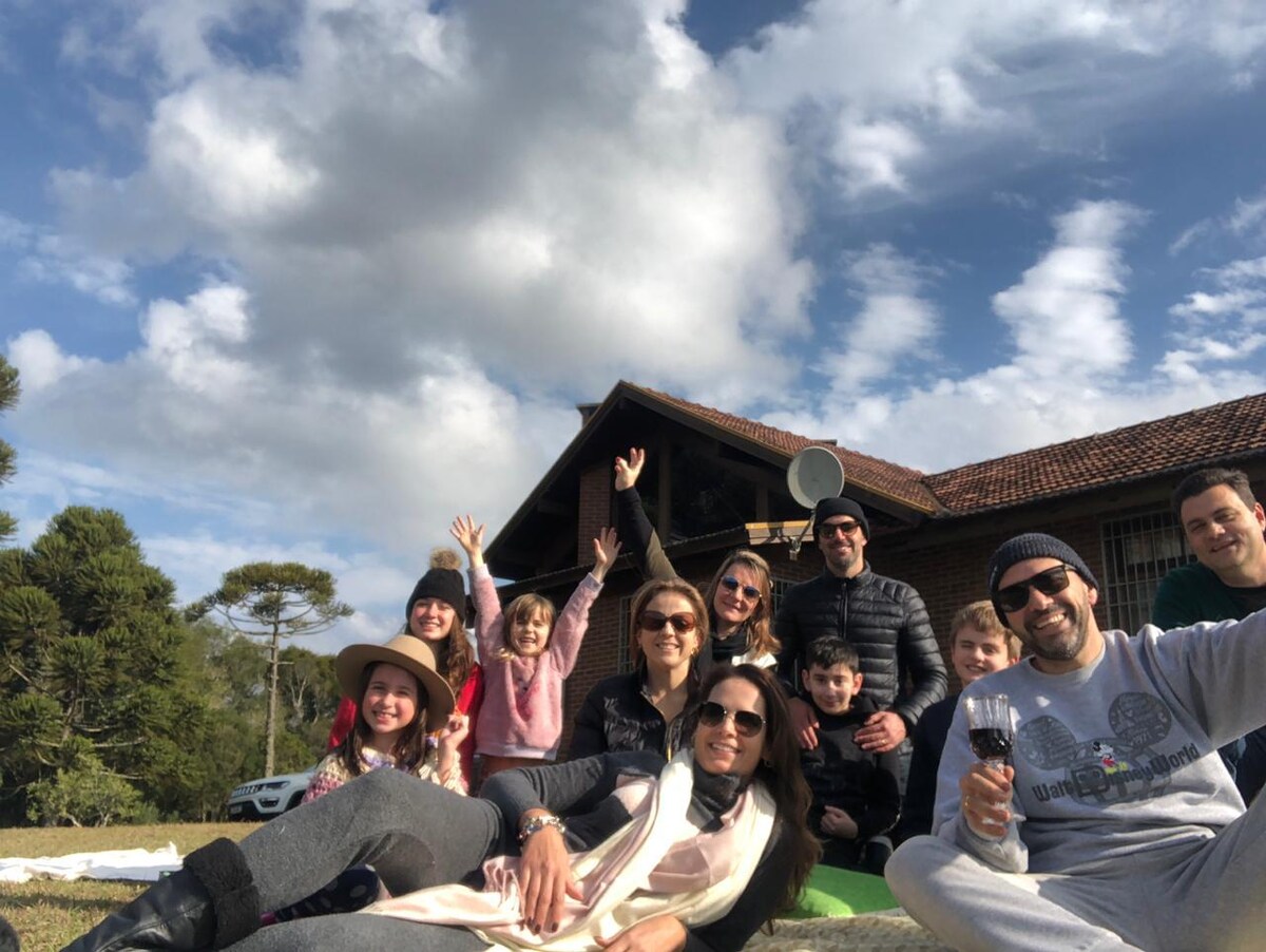A group of people is gathered on green grass in front of a rustic house with a tiled roof, smiling and enjoying their time together. Trees and clouds fill the background, creating a relaxed outdoor setting. The atmosphere suggests a shared moment of joy.