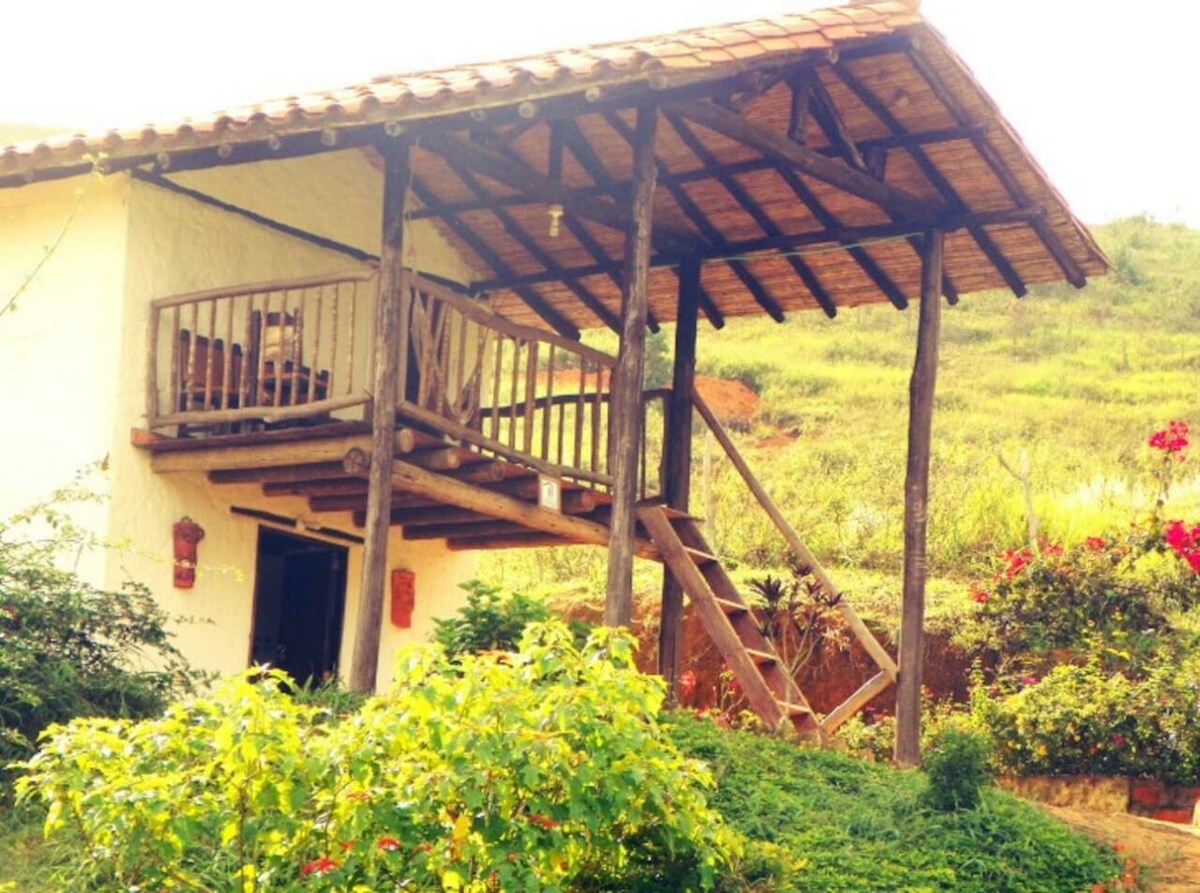 A charming cabin is depicted with a wooden balcony, featuring steps leading up to it. Surrounding greenery includes vibrant shrubs and plants, while a grassy hill rises gently in the background, enhancing the serene rural setting.
