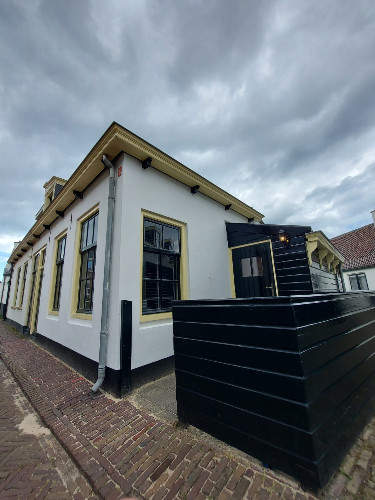The exterior of a charming cottage is shown, featuring a white facade with dark trim and a small black entranceway. Large windows allow for ample natural light, and a brick pathway leads up to the front door. The sky is overcast, adding a moody atmosphere.