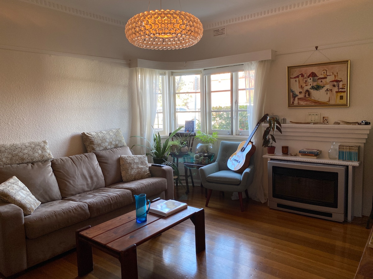 A comfortable living room is presented, featuring a light brown sofa and a cozy armchair beside a wooden coffee table. Natural light fills the space through large windows, surrounded by greenery and decorative items. A textured chandelier hangs from the ceiling, adding an element of style.