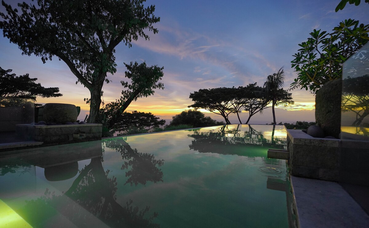 An infinity pool reflects the vibrant hues of a sunset, framed by lush tropical trees. The tranquil water appears to blend seamlessly with the sky, creating a serene atmosphere, while the surrounding landscape showcases the natural beauty of the hillside setting.