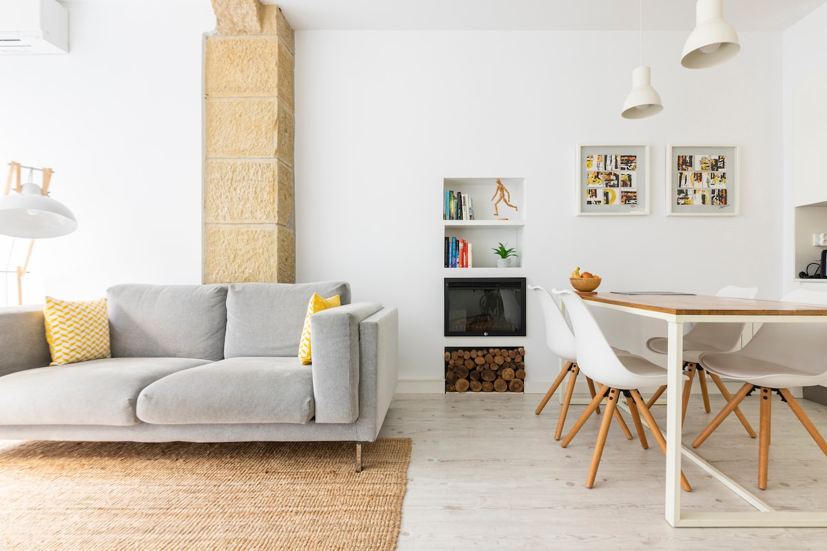 A modern living area features a light grey sofa adorned with yellow accent pillows. A wooden bookshelf displays books and decorative items, while a cozy dining table with white chairs is set nearby. Natural light streams through the space, enhancing the clean design.