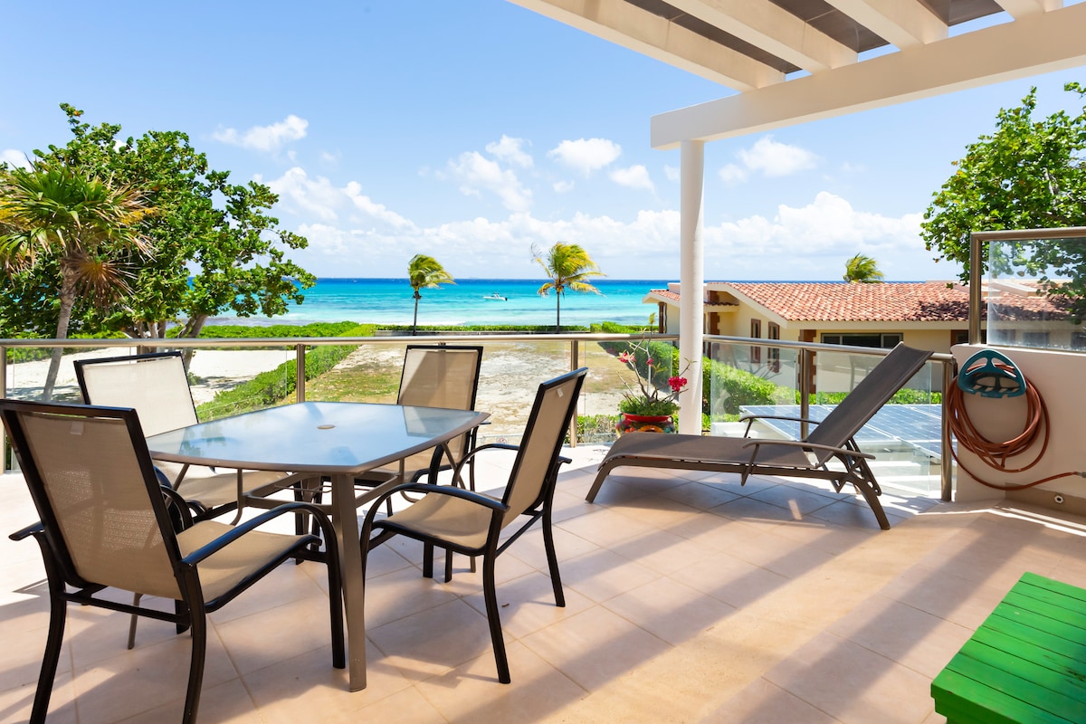 An expanded patio offers breathtaking ocean views, furnished with a glass-top dining table and six chairs. A reclining lounge chair is positioned nearby. Lush greenery and palm trees border the patio, while the vibrant blue waters of the Caribbean Sea create a serene backdrop.
