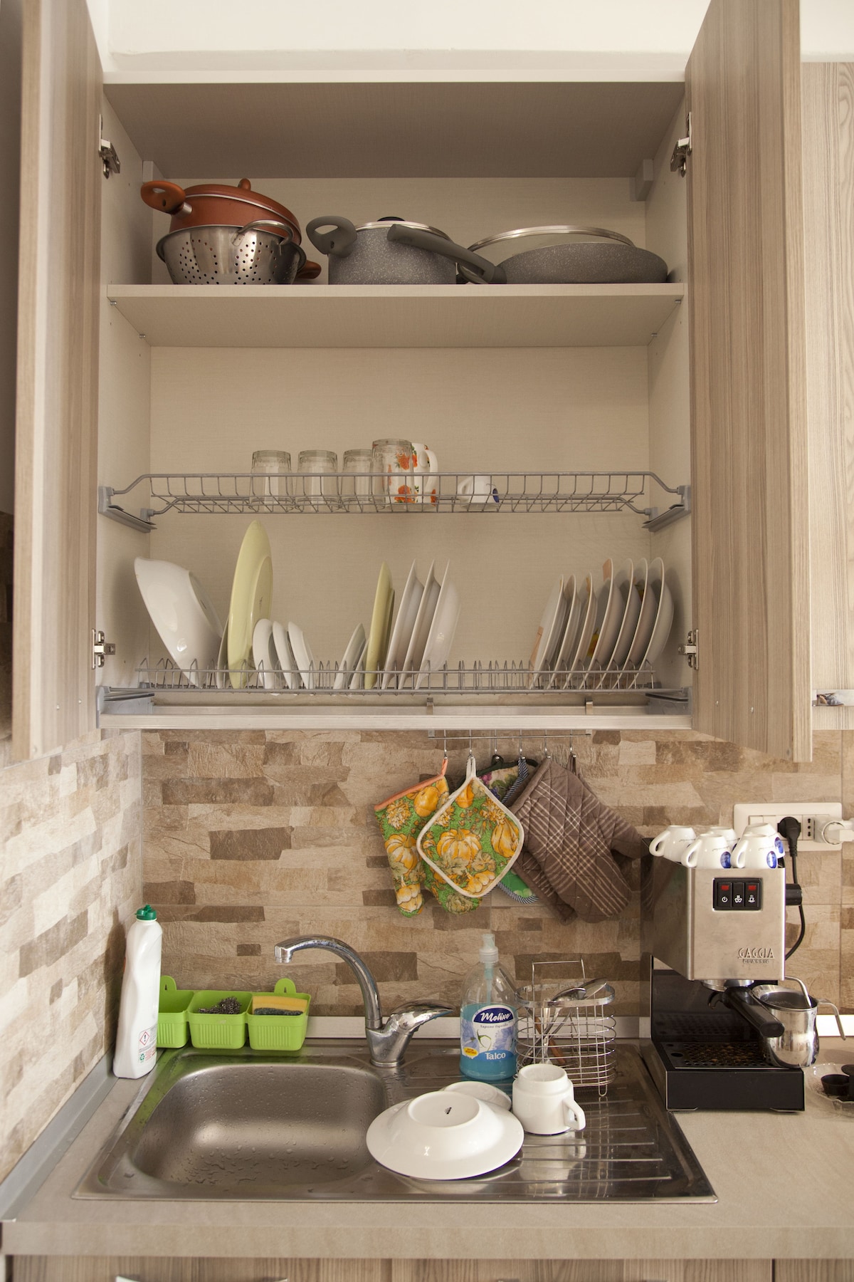 The kitchen area features a neatly organized cabinet containing various cooking pots and plates. Glassware is arranged on the middle shelf, while a drying rack holds clean dishes. A sink with dishwashing liquid and a coffee machine are visible, along with colorful kitchen towels hanging nearby.