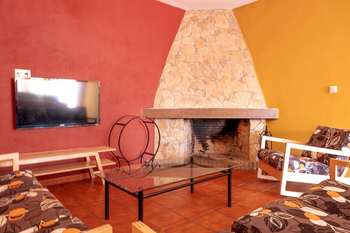 A cozy living area features a stone fireplace, complemented by a flat-screen TV mounted on the wall. Two patterned sofas are arranged around a glass coffee table, offering a welcoming space for relaxation. The walls are painted in warm hues of red and yellow.