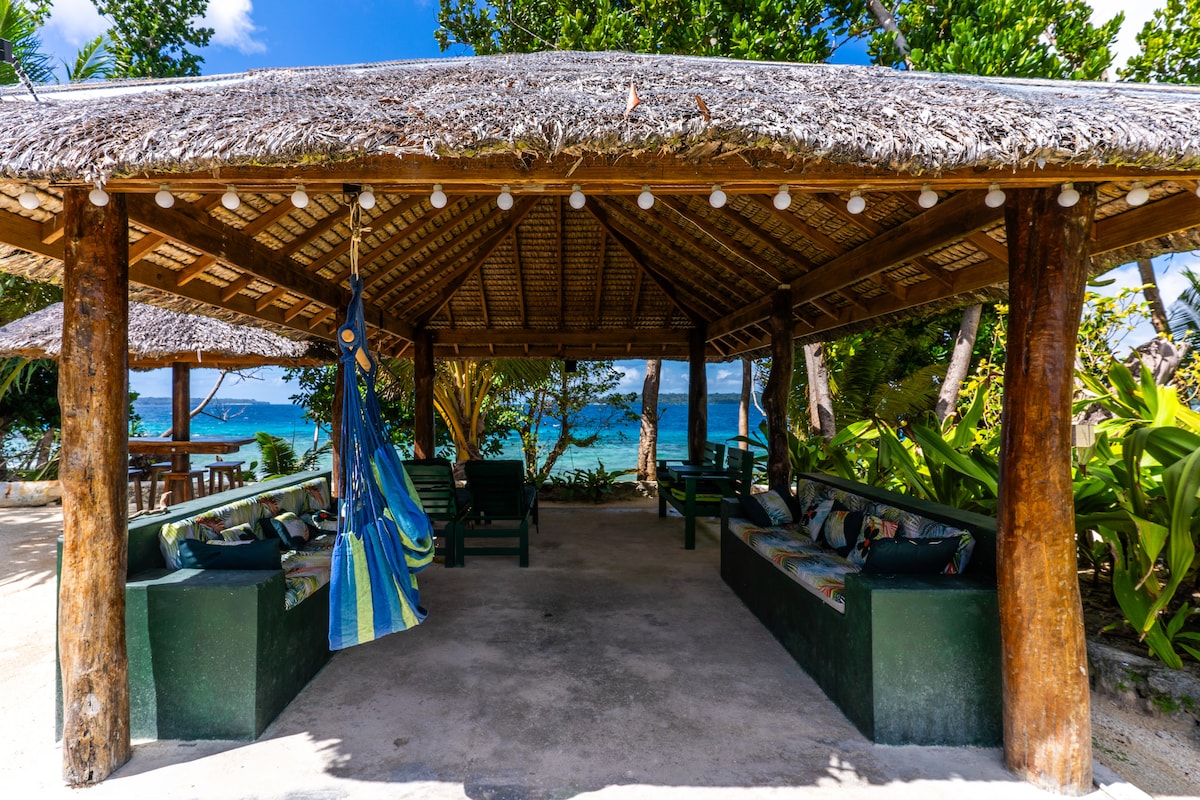 A covered outdoor area features wooden beams and a thatched roof, providing shade and comfort. Seating is arranged along the sides, with cushions enhancing relaxation. A hammock hangs nearby, inviting rest. The blue waters and greenery are visible in the background, creating a tranquil setting.