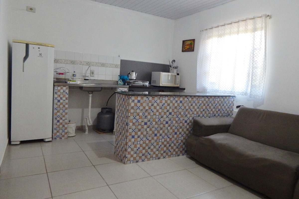 A well-lit kitchen area features a compact refrigerator and a microwave atop a tiled countertop. A cozy sofa sits nearby, with a window draped in light-colored curtains allowing natural light to enter the space. Decorative art piece is mounted on the wall.