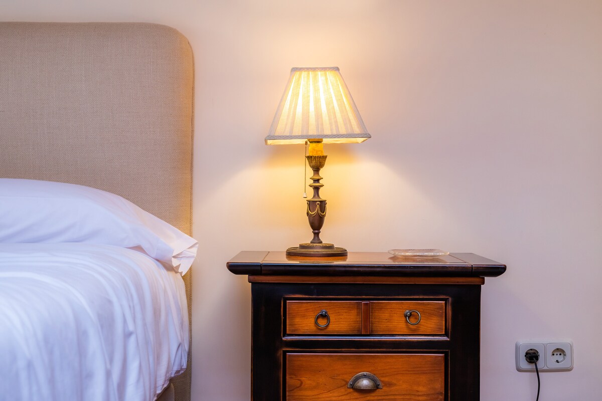 A cozy bedside area is featured, with a lamp casting a warm glow on a wooden nightstand. The spacious bed is dressed in crisp white linens, creating a serene atmosphere. The wall displays a soft hue, complementing the elegant furnishings.