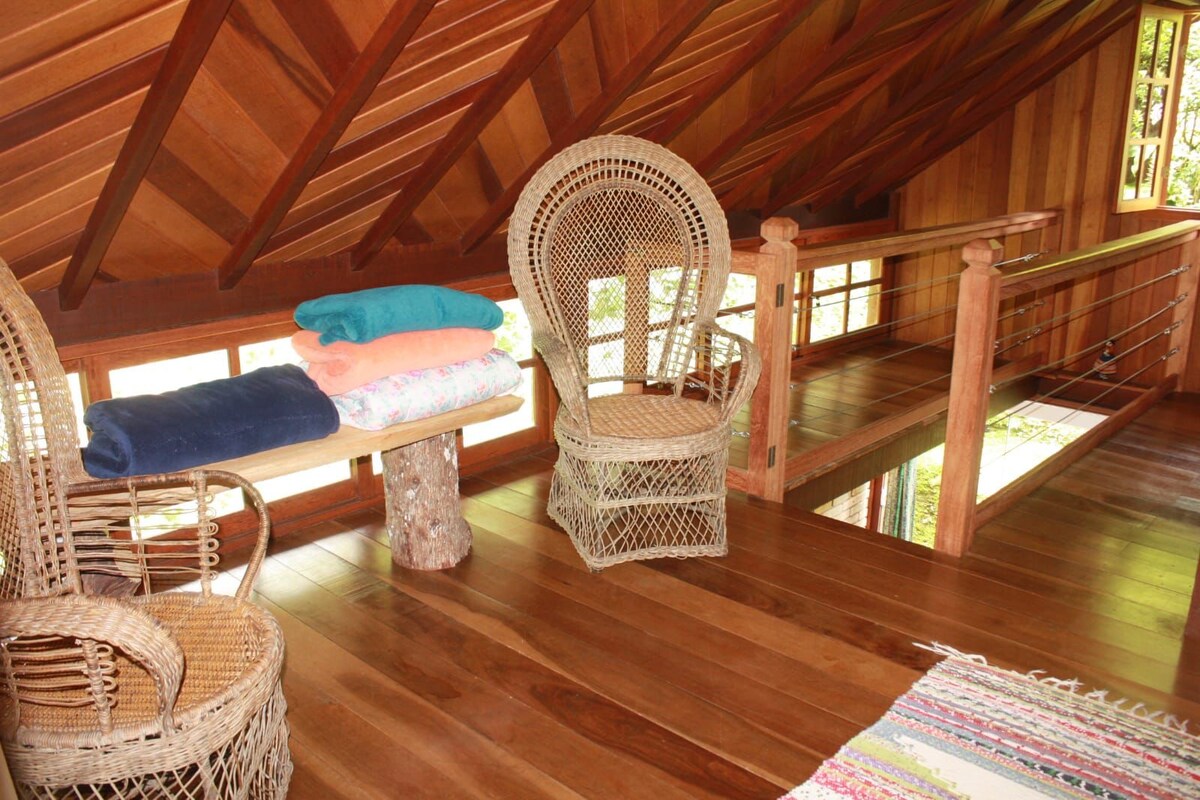 An inviting loft area features two woven chairs positioned beside a wooden table with stacked towels. Natural wood accents create warmth, while large windows allow ample light and views of the surrounding greenery. A colorful area rug adds a touch of comfort to the wooden floor.