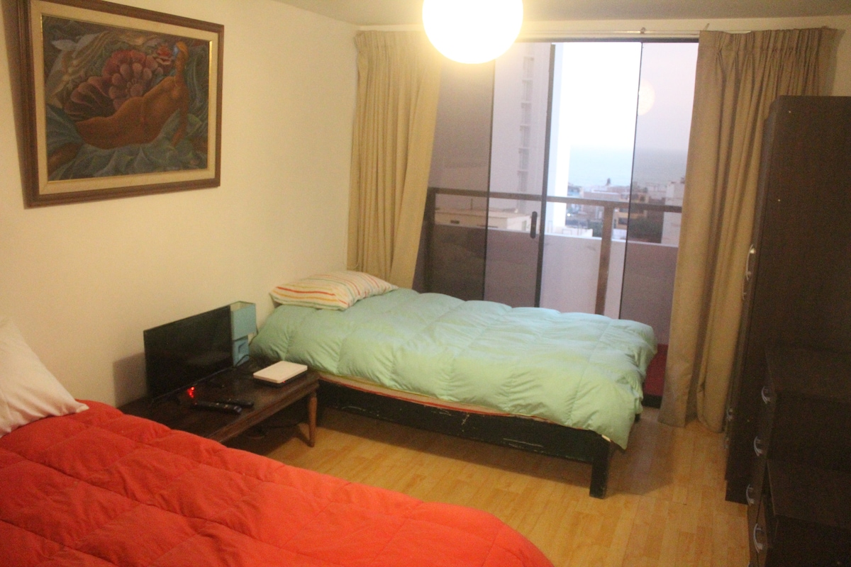 A bright sleeping area is furnished with two beds covered in colorful bedding. A wall-mounted television is placed on a low table, and a large window offers sea views. Soft curtains frame the window, allowing for natural light to illuminate the space.
