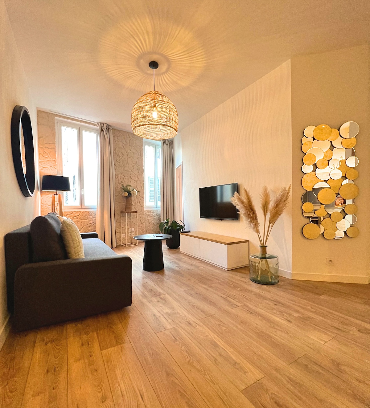 A modern living area features a textured wall and a large window with sheer curtains. A dark sofa is positioned next to a small black table. A tall lamp and potted plants add greenery. A circular mirror reflects ambient light, enhancing the space.