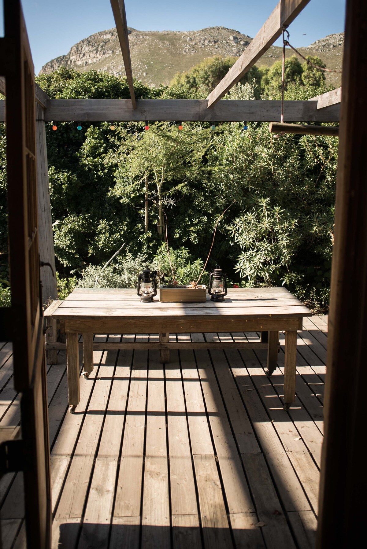 A wooden outdoor table is set on a spacious deck, surrounded by lush greenery. Lanterns are placed on the table, casting gentle shadows across the wooden surface. The view is framed by an open doorway, leading to a serene and natural setting.