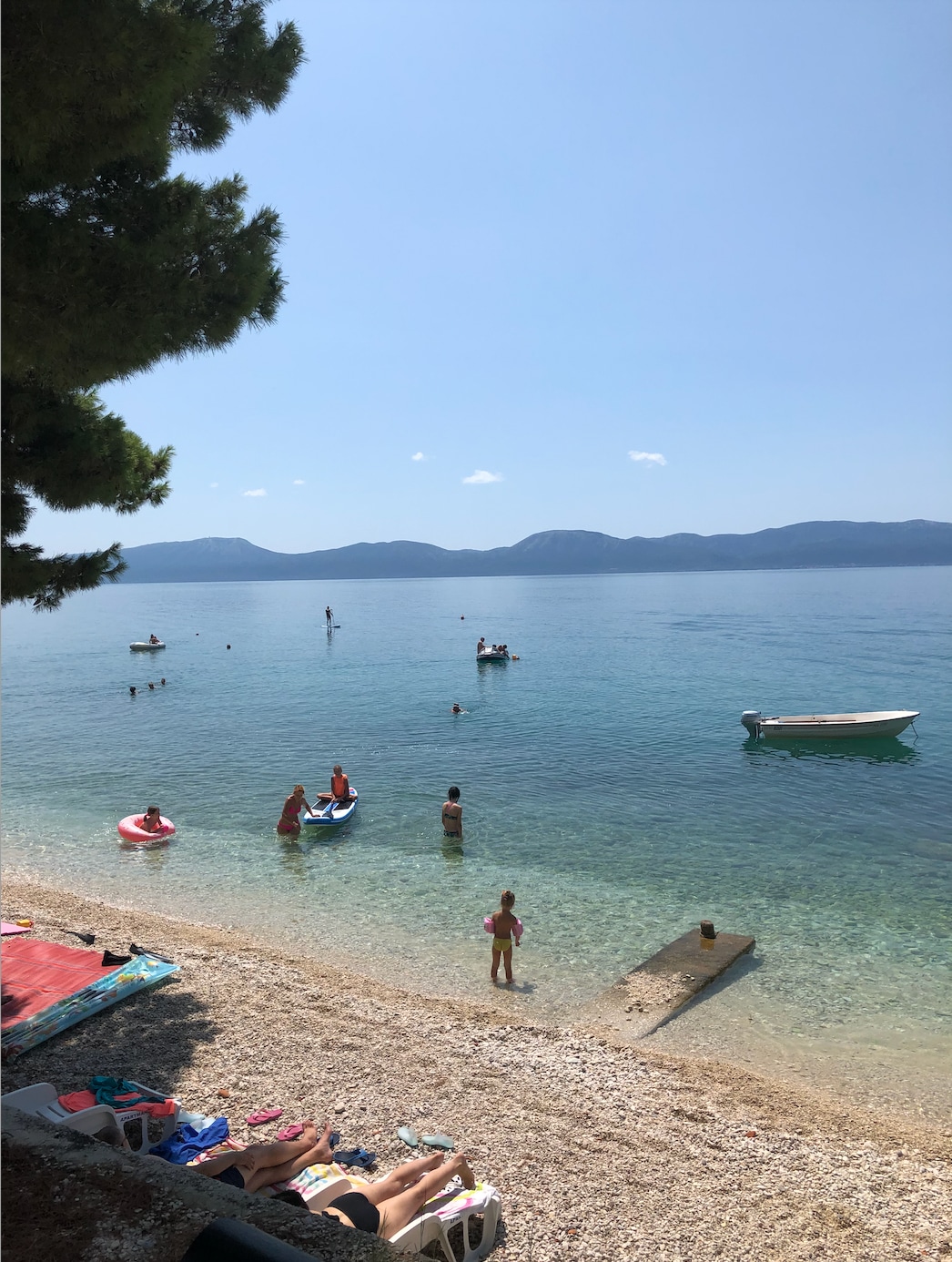 A calm beach scene showcases clear blue waters with several swimmers and boats visible in the distance. Soft sand meets the gentle waves, while hills rise in the background under a bright sky. A mix of activities can be observed, inviting relaxation and enjoyment.