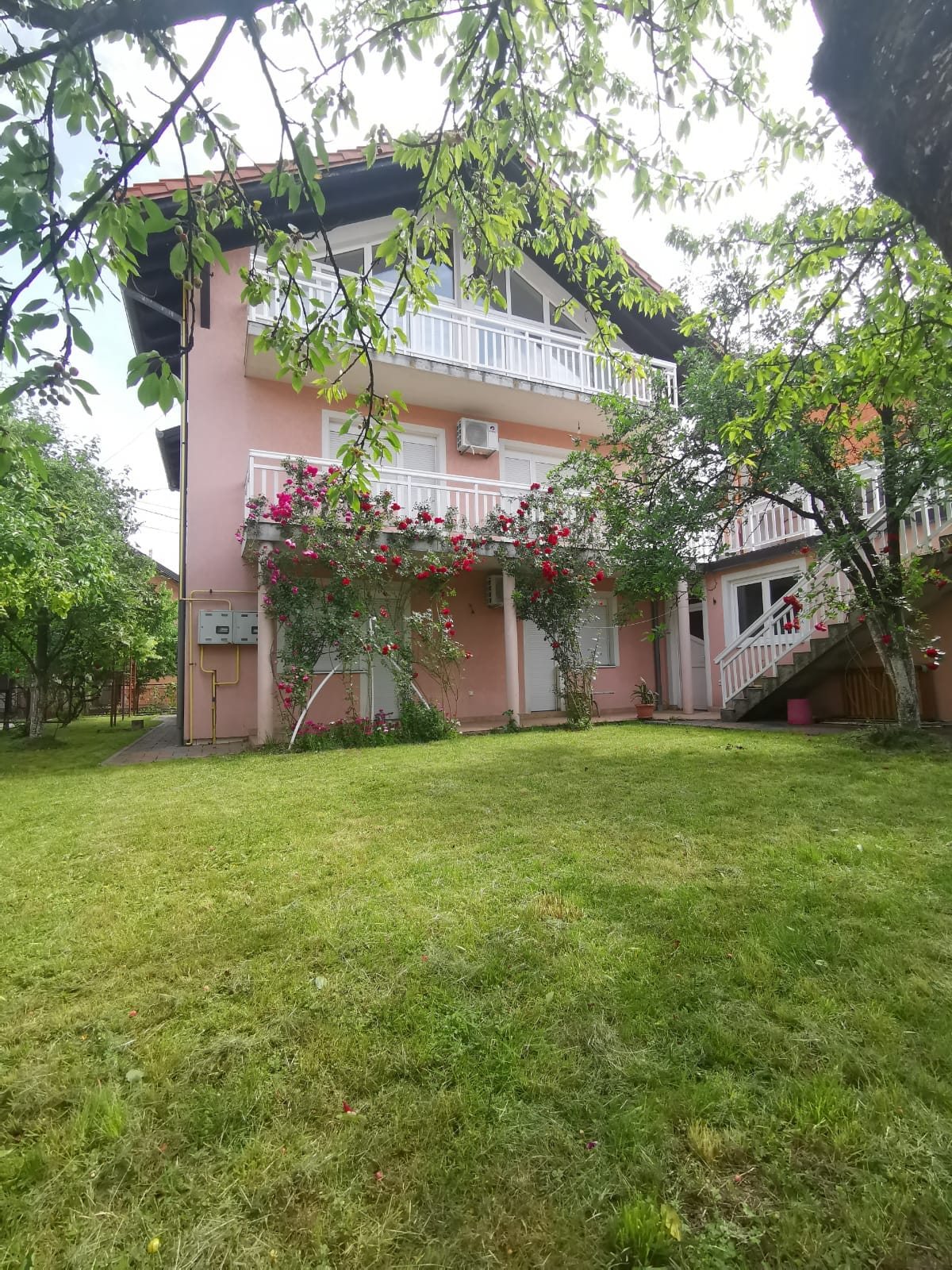 The exterior of a pink two-story house is surrounded by lush greenery and flower-covered trellises. An inviting garden area features well-maintained grass, and multiple balconies overlook the yard, offering a serene outdoor space.