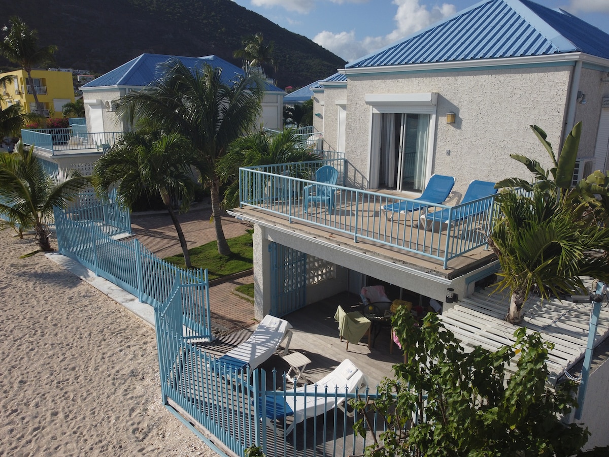The two-story villa is situated on a sandy beach, featuring a wraparound deck with blue lounge chairs. A private garden is visible, bordered by palm trees. The light-colored exterior contrasts with the vibrant blue roof, and large glass doors provide access to the indoor living space.