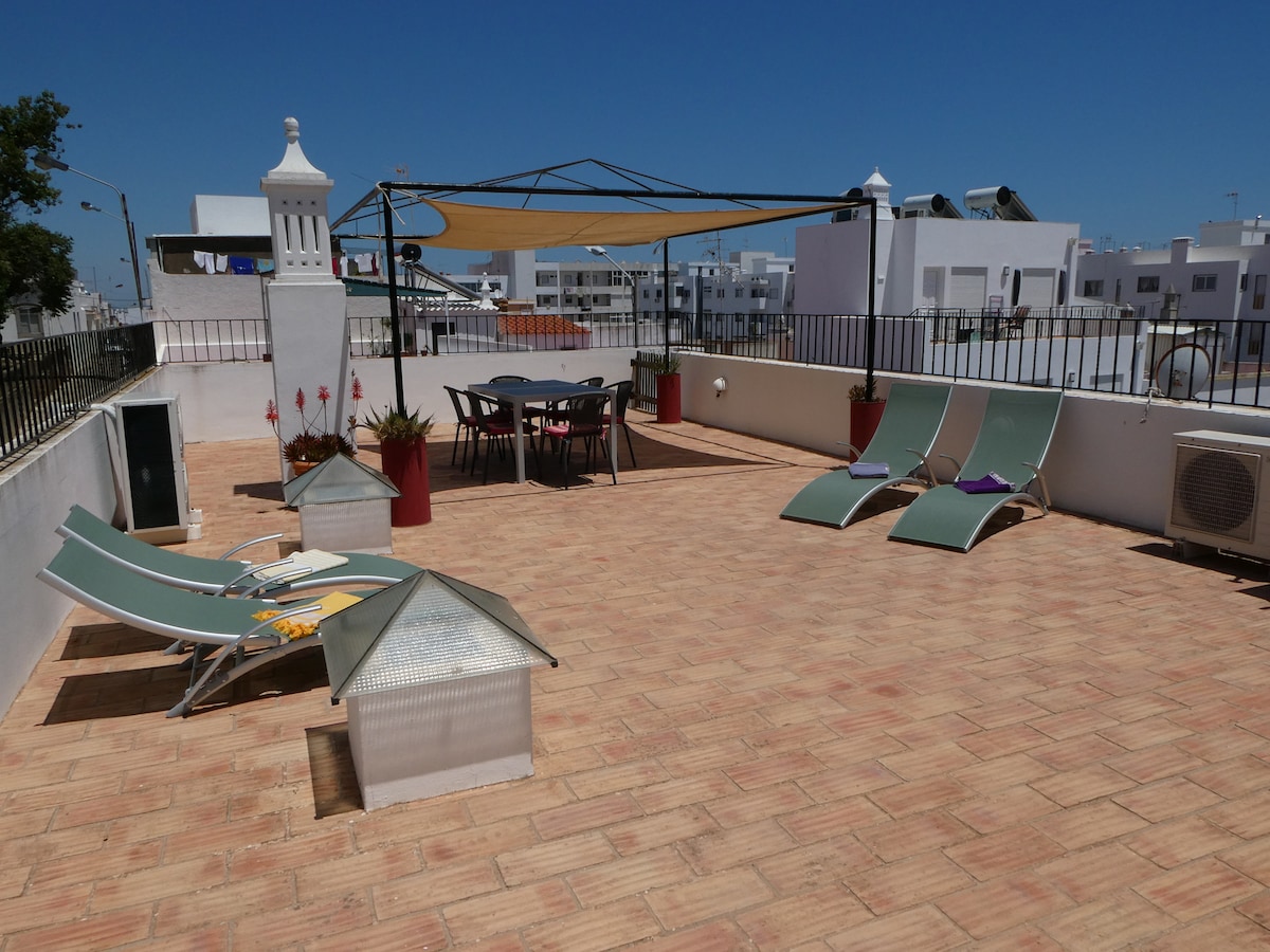 The rooftop terrace features a spacious area with sun loungers and a shaded dining table. Potted plants add a touch of greenery, while the surrounding railing ensures safety. The sun shines brightly, creating an open and airy atmosphere with views of the surrounding area.