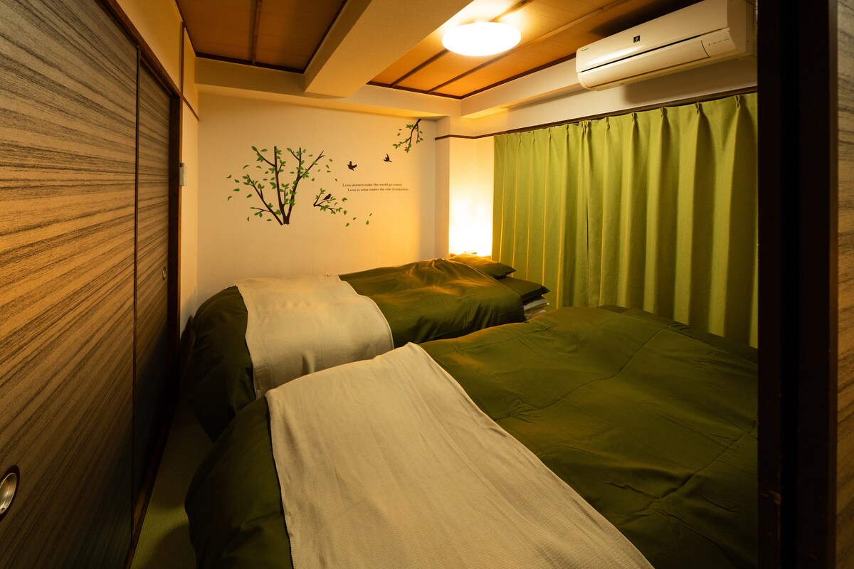 A comfortably arranged room features two spacious green beds adorned with matching linens. A soft light is provided by a lamp on the side, and a wall with nature-themed artwork creates a serene ambiance. Green curtains add privacy and warmth.