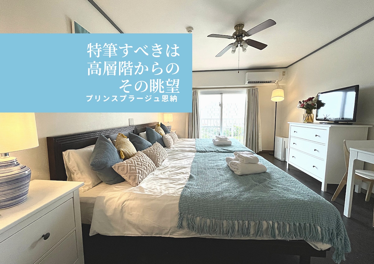The room features two double beds with fresh linens, a ceiling fan, and a TV unit. Large windows provide natural light and offer a view of the surroundings. A white dresser and a decorative vase add to the room's functionality and style.