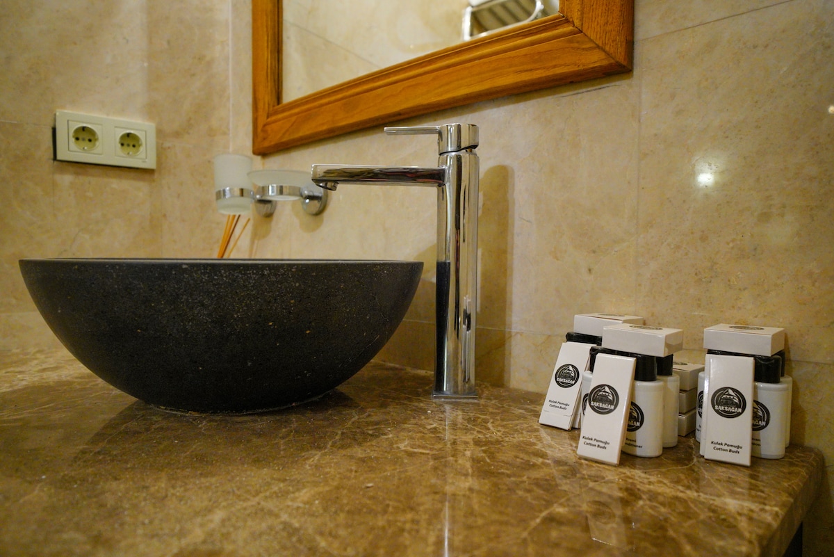 A stylish bathroom sink is showcased with a sleek, dark stone bowl. A modern faucet is positioned beside a wooden-framed mirror. Several compact toiletries are neatly arranged on the marble countertop, complementing the elegant design and cleanliness of the space.