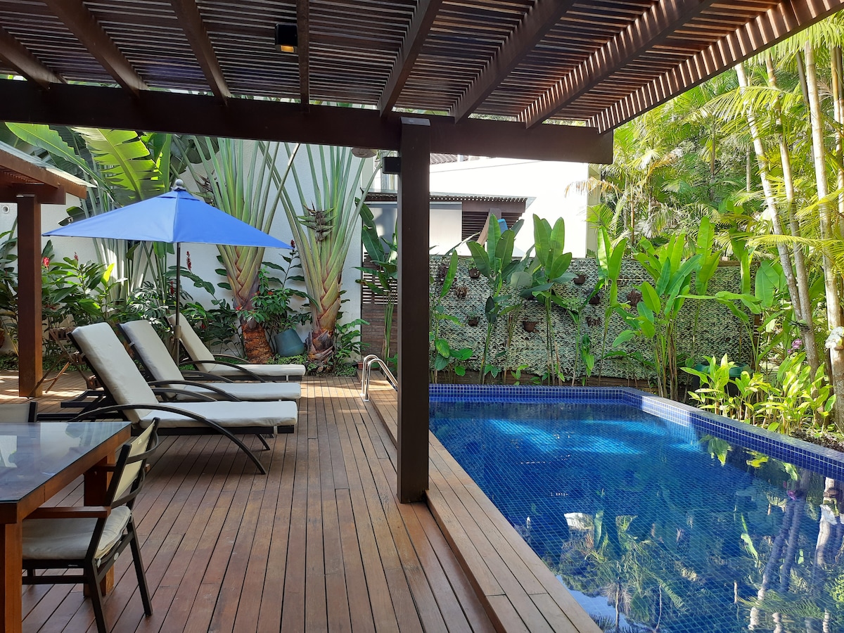 An outdoor area features a refreshing pool surrounded by lush tropical plants. Deck chairs are positioned near the pool under a large blue umbrella. Wood decking provides a warm contrast to the water's blue tones, enhancing the overall tranquil ambiance.