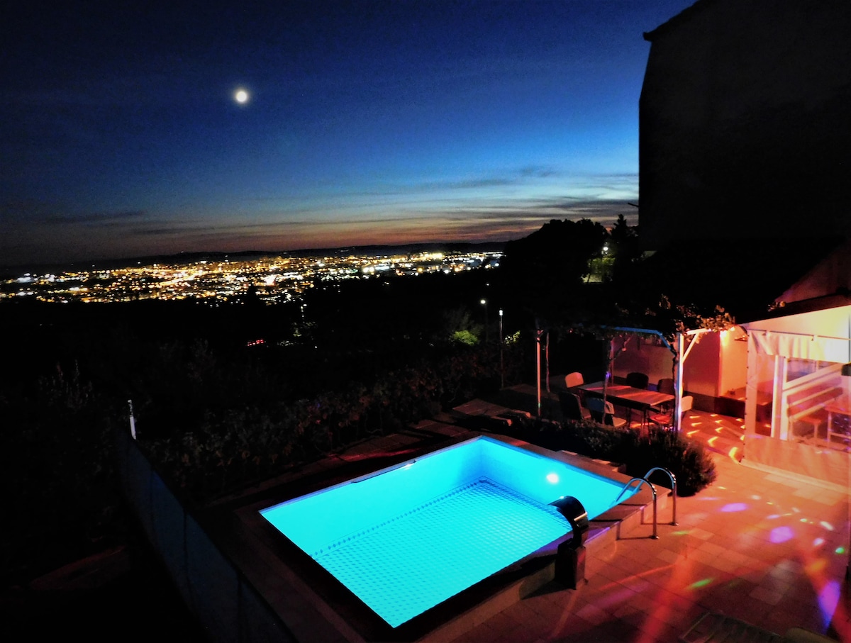 The heated swimming pool is illuminated in soft blue light, surrounded by scenic views of the city below. The evening sky transitions from sunset colors to deep blue, with stars beginning to appear. A patio area with seating is visible, enhancing the outdoor ambiance.