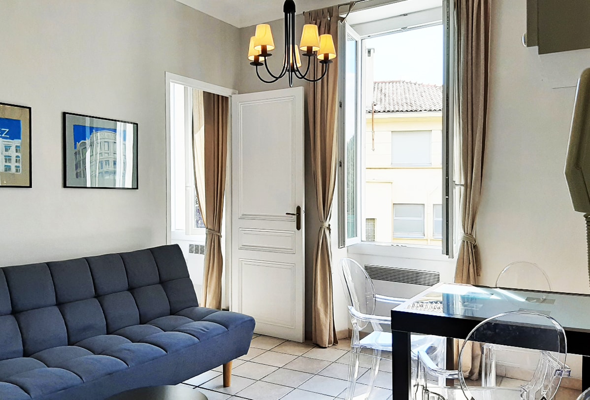 The living area features a comfortable sofa bed in a soft gray hue, paired with a clear dining table. Natural light floods the space through large windows adorned with light curtains, while a modern chandelier hangs above, creating a bright and airy atmosphere.