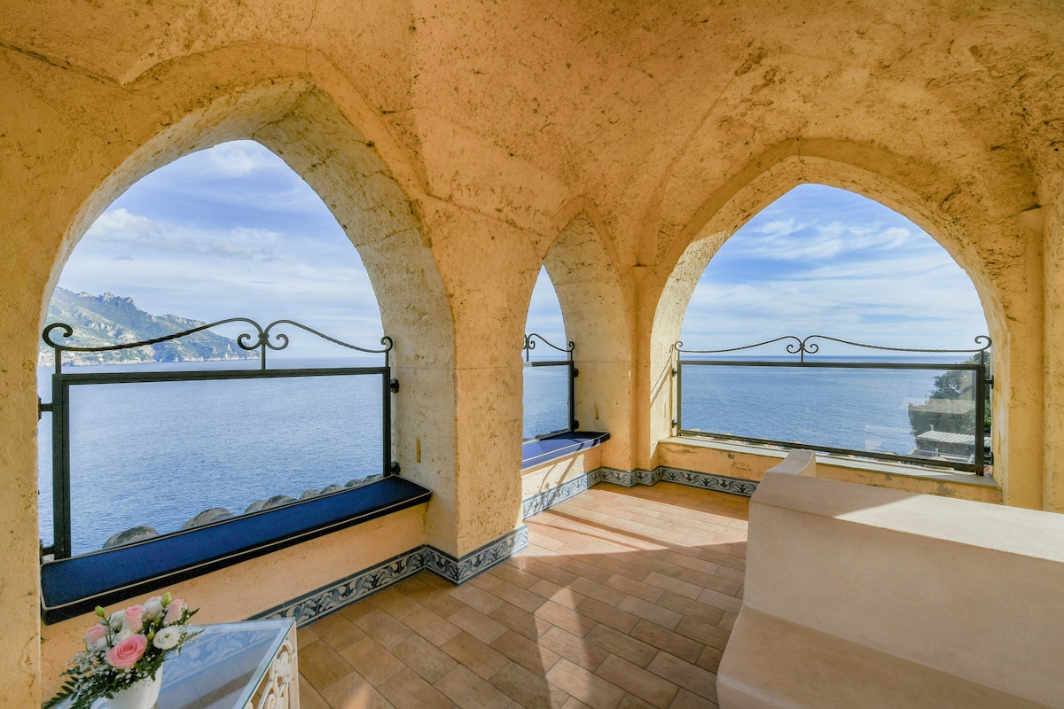 A sunlit terrace showcases three arched windows with wrought iron railings, offering sweeping views of the sea and coastline. Soft seating areas are present along the windowsills, and decorative tiles adorn the floor, creating a serene space for relaxation.
