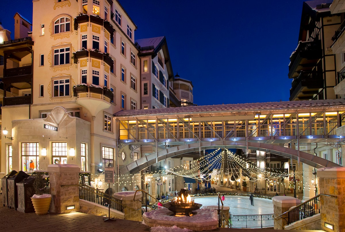 A scenic view of a charming outdoor plaza at night, featuring a large skating rink surrounded by elegant buildings. Light strings create a warm ambiance, and a circular fire pit invites guests to gather. The architecture displays a blend of traditional and modern design elements.