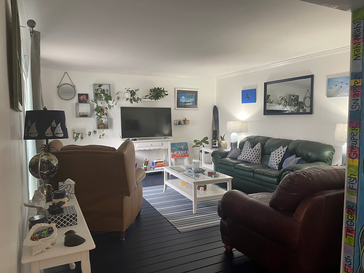 The living room features a combination of comfortable seating, including a brown leather sofa and a green leather couch. A coffee table is centered in the space, surrounded by decorative plants and coastal-themed artwork. Natural light fills the room through large windows.