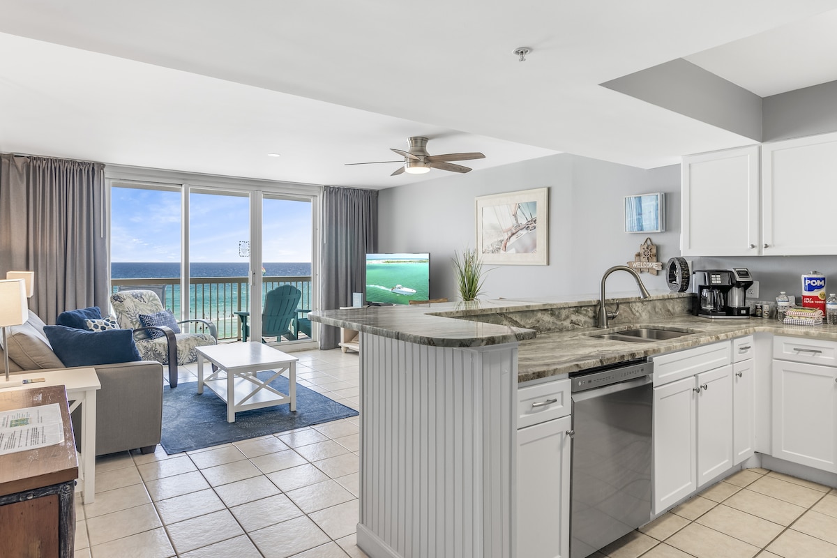 A spacious living area features a bright open layout, with a modern kitchen showing stainless steel appliances and granite countertops. Comfortably furnished, the space includes a cozy sofa facing large glass doors, leading to a private balcony with stunning ocean views.