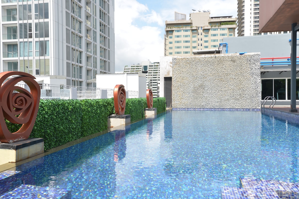 A stylish rooftop pool is surrounded by modern decorative features and lush greenery. The clear water reflects the nearby buildings, creating a serene atmosphere. Sunbeds line the pool area, inviting relaxation under the bright sky.