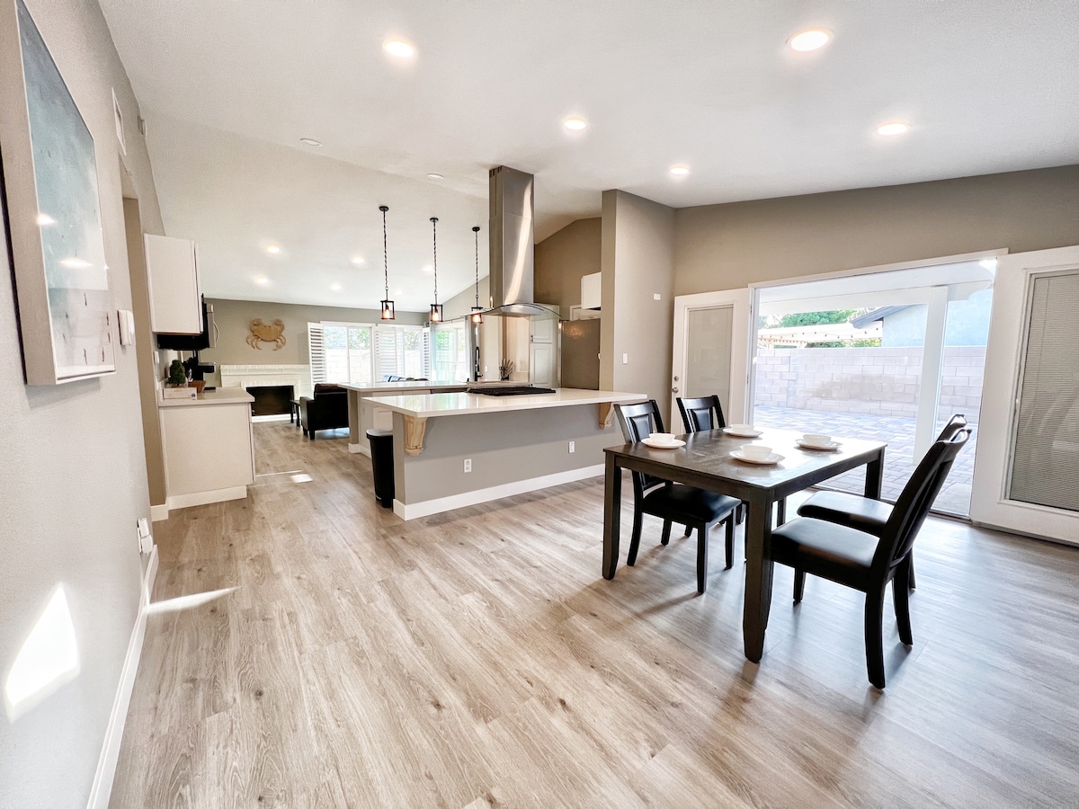 A contemporary dining area features a dark wooden table surrounded by four chairs. Open concept architecture is highlighted, allowing natural light to enter through sliding glass doors. The kitchen, with modern appliances and a large island, is visible in the background.