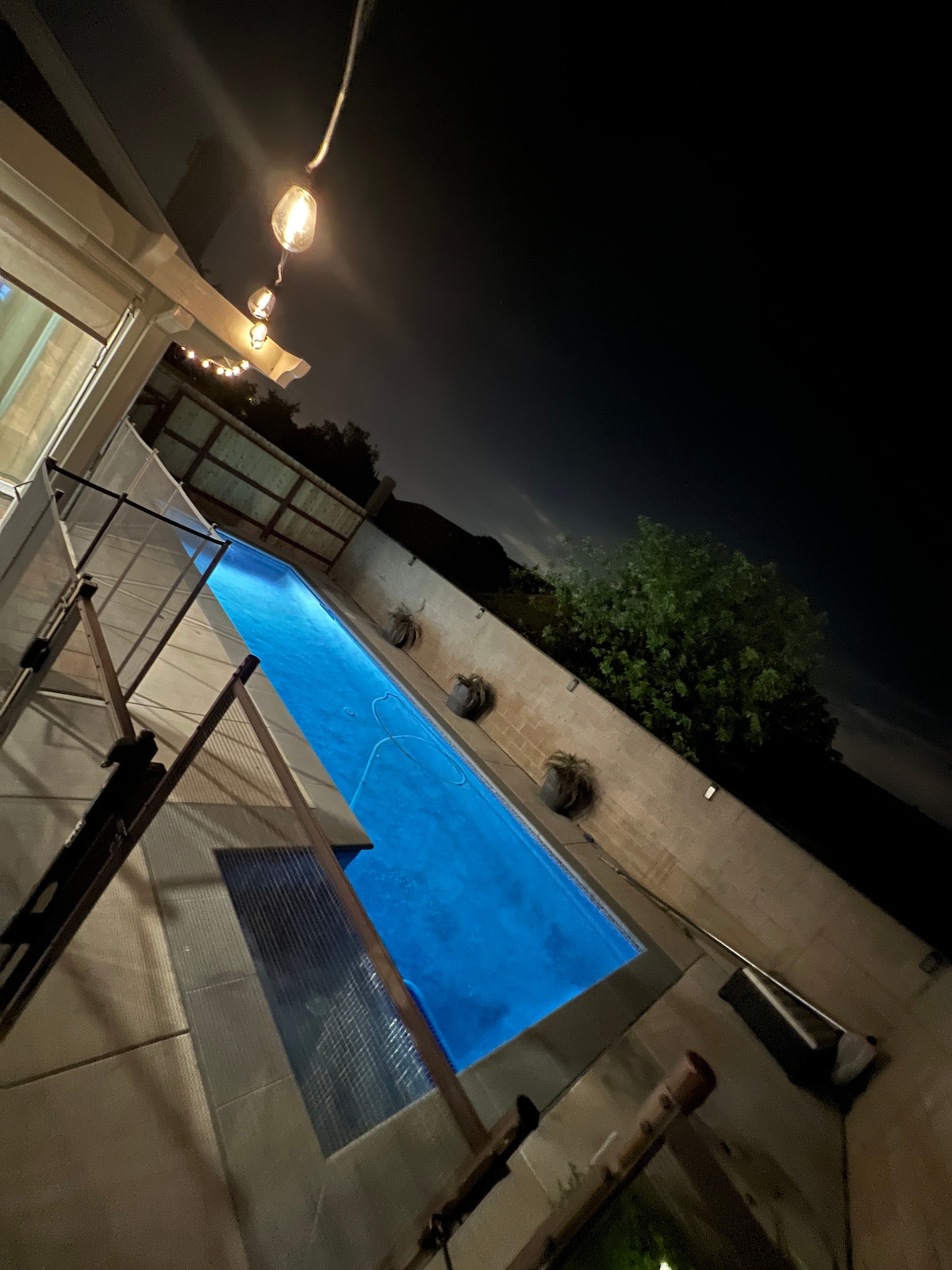 The private pool is illuminated under the night sky, showcasing its clear blue water. Surrounding the pool, safety fencing ensures a secure environment. Soft lighting hangs above, creating a warm ambiance against the dark backdrop. The tranquil space offers a peaceful retreat.