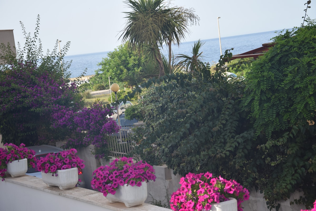 A vibrant display of blooming flowers in pink and purple hues is seen in planter boxes along the terrace. Lush greenery surrounds the area, complementing the view of the sea in the background, offering a serene outdoor space.