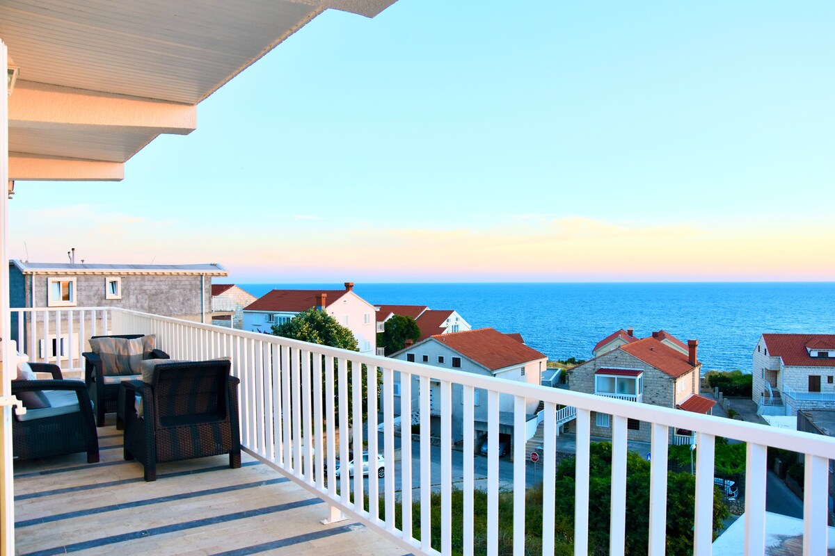 A balcony is presented with comfortable seating arrangements overlooking a sweeping view of the blue Adriatic Sea. Residential buildings with red roofs are visible in the foreground, while a clear sky transitions softly from light blue to a warm hue.
